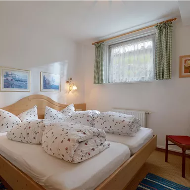 Ein gemütliches Schlafzimmer mit einem Doppelbett und vielen Kissen. Es gibt ein Fenster mit Vorhängen und einen Stuhl in der Ecke.
