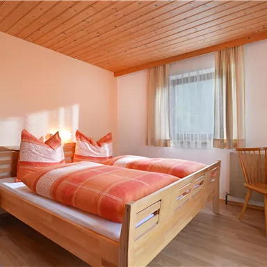 Ein gemütliches Schlafzimmer mit zwei Betten und orangefarbenen Kissen. Der Raum hat Holzdecken und ein Fenster, das viel Tageslicht hereinlässt.
