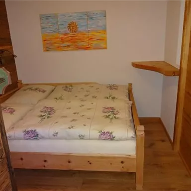 Ein gemütliches Schlafzimmer mit einem Holzbett und einer Matratze, die mit Blumenmuster bezogen ist. An der Wand hängt ein farbenfrohes Bild, und es gibt einen kleinen Ablagetisch.