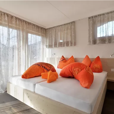 Ein gemütliches Schlafzimmer mit einem großen Bett und vielen orangefarbenen Kissen. Die Fenster lassen viel Licht herein und die Vorhänge verleihen eine warme Atmosphäre.