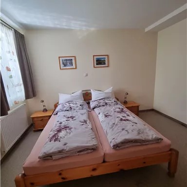 Ein gemütliches Schlafzimmer mit einem Doppelbett und zwei Nachttischen. Die Wände sind hell und es gibt Fenster, die natürliches Licht hereinlassen.