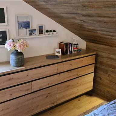 A cozy bedroom with a wooden dresser and decorative pictures on the wall. On the nightstand, there are flowers and books, while warm light shines from a nook.
