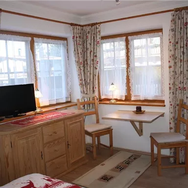 Ein gemütliches Zimmer mit Holzdekor, einem Fernseher und einem kleinen Tisch. Große Fenster lassen viel Licht herein und es gibt Stühle für Gäste.