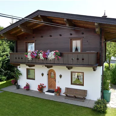 Ein gemütliches Haus mit einem oberen Balkon, geschmückt mit bunten Blumen. Der Garten ist grün und einladend, umgeben von Bäumen.