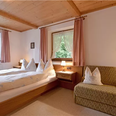 Ein gemütliches Schlafzimmer mit zwei Betten und einem kleinen Sitzbereich. Große Fenster lassen viel Tageslicht herein und bieten einen Blick ins Grüne.