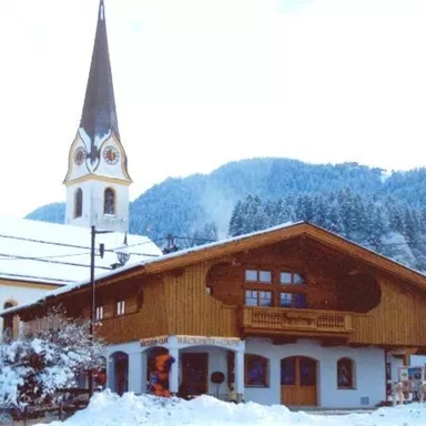 A picturesque winter landscape with a traditional wooden building and a church in the background. The surroundings are covered with snow and surrounded by mountains.