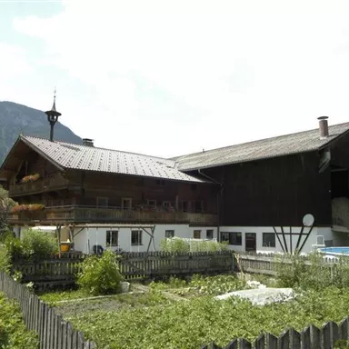 A traditional alpine house with two buildings and a well-kept garden. In the background, mountains and a clear sky can be seen.