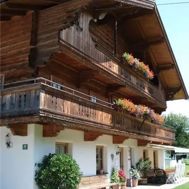 A traditional wooden house with two balconies and colorful flowers. The facade is bright and inviting, surrounded by a well-maintained garden.