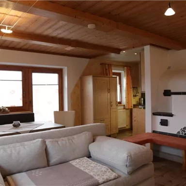 Ein gemütlicher Wohnraum mit einem großen Sofa und einem Ofen. Die Holzbalkendecke und die Fenster sorgen für eine warme Atmosphäre.