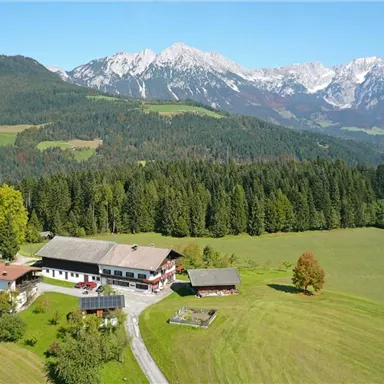 Eine idyllische Landschaft mit einem Bauernhaus und grünen Wiesen. Im Hintergrund erheben sich majestätische Berge unter einem blauen Himmel.