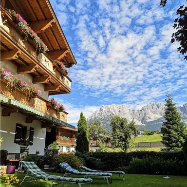 Ein malerisches Chalet mit blühenden Balkonen und Liegen im Garten. Im Hintergrund erheben sich majestätische Berge unter einem blauen Himmel.