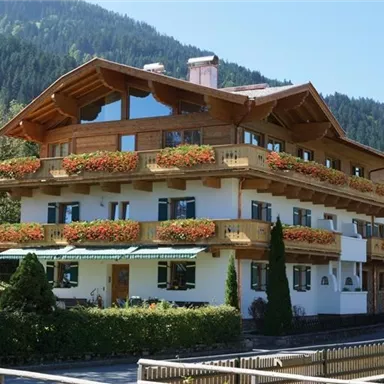 Ein traditionelles Haus im Alpenstil mit Balkonen, die mit bunten Blumen geschmückt sind. Im Hintergrund sind grüne Berge zu sehen.
