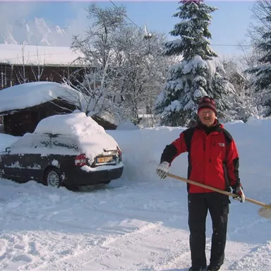 Ein Mann steht im Schnee und hält eine Schneeschaufel. Im Hintergrund sind verschneite Bäume und ein mit Schnee bedecktes Auto zu sehen.