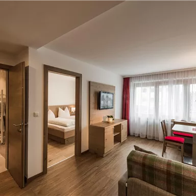 A modern living kitchen with a cozy table and a sofa. In the background, a bedroom and a bathroom are visible.