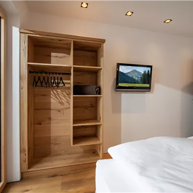 Ein modernes Schlafzimmer mit einem Holzschrank und einem kleinen Fernseher an der Wand. Große Fenster lassen viel Tageslicht herein und bieten einen Blick auf die Umgebung.
