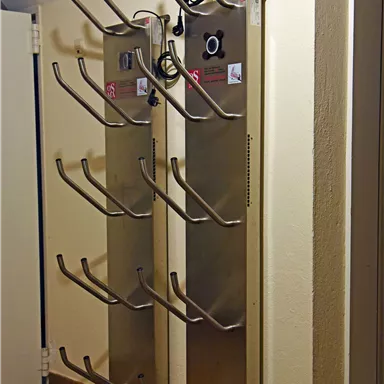 A stainless steel coat rack with several hooks for jackets or bags. The wall is light and the floor is made of wood.