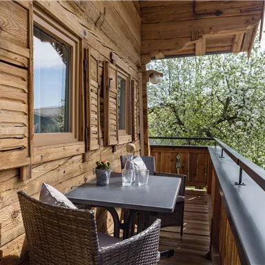Ein gemütlicher Balkon aus Holz mit einem Tisch und Stühlen. Im Hintergrund blühen Bäume und der Himmel ist klar.