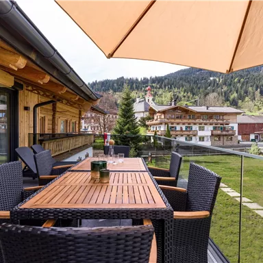 Ein eleganter Balkon mit einem Holztisch und Stühlen aus Rattan. Im Hintergrund sind malerische Berge und Holzchalets zu sehen.