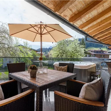 Ein gemütlicher Balkon mit einem Tisch und Stühlen. Im Hintergrund sind Bäume und Berge zu sehen.