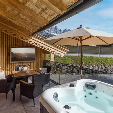Ein gemütlicher Außenbereich mit einem Whirlpool und einem Esstisch unter einem Sonnenschirm. Im Hintergrund sind die Berge und ein klarer Himmel sichtbar.