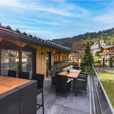 Eine moderne Terrasse mit Holzmöbeln und Blick auf grüne Wiesen und Berge. Die Umgebung ist ruhig und einladend.