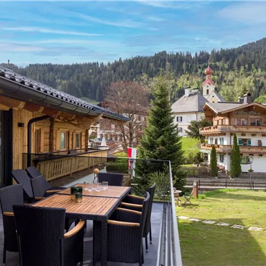 Eine gemütliche Terrasse mit einem Holztisch und Stühlen, umgeben von einer grünen Wiese und Häusern in traditioneller Architektur. Im Hintergrund sind Bäume und majestätische Berge zu sehen.
