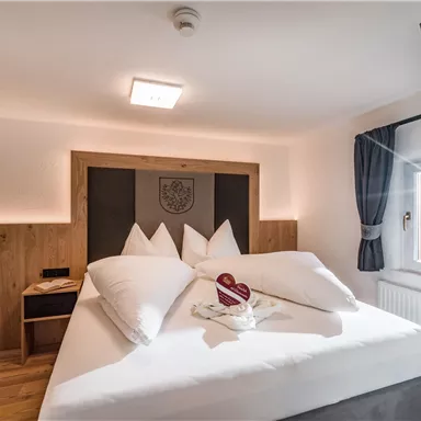 Ein gemütliches Zimmer mit einem großen Bett und modernen Möbeln. Hell erleuchtet und freundlich gestaltet, mit Fenstern, die Tageslicht hereinlassen.