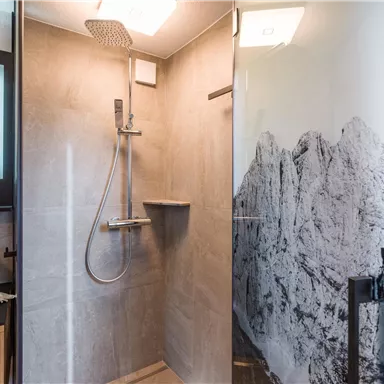 Eine moderne Dusche mit grauen Fliesen und einer gläsernen Abtrennung. An der Wand ist ein traumhaftes Bergmotiv zu sehen.