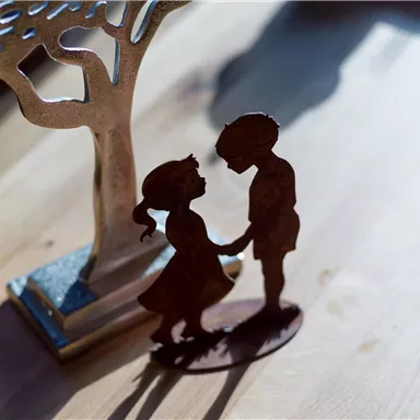 Eine filigrane Metallfigur, die ein Kindermädchen und einen Jungen zeigt, die sich die Hände halten. Im Hintergrund steht ein verzierter Baumständer.