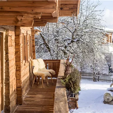 Ein Holzhaus mit einer verzierten Veranda, umgeben von schneebedeckten Bäumen. Die ruhige Winterlandschaft vermittelt eine gemütliche Atmosphäre.