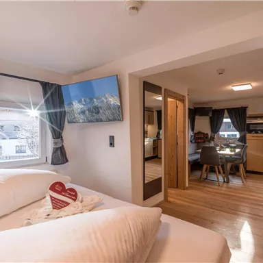 Ein gemütliches Schlafzimmer mit zwei Betten, einem Fernseher und großen Fenstern. Der angrenzende Wohnbereich ist modern und hell eingerichtet.