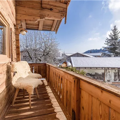 Ein gemütlicher Balkon mit Holzgeländer und zwei bequemen Stühlen. Im Hintergrund sind schneebedeckte Bäume und ein klarer Himmel zu sehen.