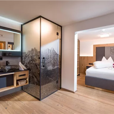 Ein modernes Hotelzimmer mit einem eleganten Badezimmer und einem gemütlichen Schlafbereich. Die Einrichtung ist stilvoll und hell, mit Holzdetails und klaren Linien.
