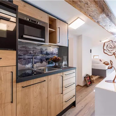 Eine moderne Küche mit Holzschränken und Edelstahlgeräten. Im Hintergrund ist ein Schlafzimmer mit einem dekorativen Rosenstandfuß zu sehen.