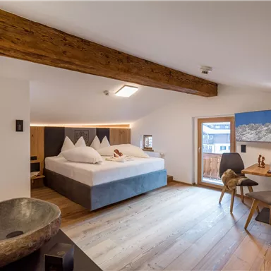 Ein modernes Zimmer mit einem großen Bett, Holzdecke und neutralen Farben. Es gibt einen gemütlichen Arbeitsplatz neben einem Fenster mit Blick auf die Berge.