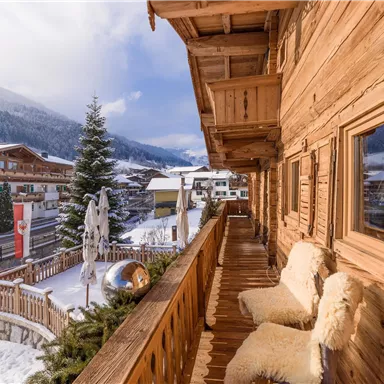 Eine gemütliche Holzveranda mit Fellkissen und Blick auf eine verschneite Landschaft. Im Hintergrund sind traditionelle chalets und ein Weihnachtsbaum zu sehen.