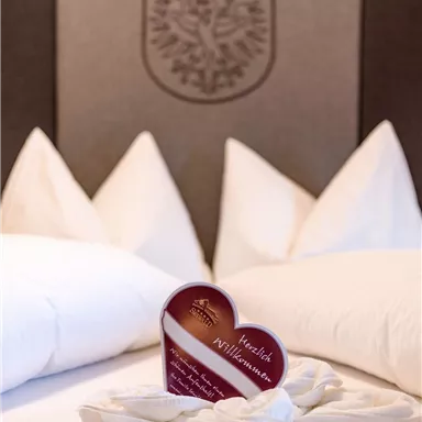 Ein gemütliches Bett mit weißen Kissen und einer herzförmigen Begrüßungskarte in der Mitte. Die Karte heißt „Herzlich Willkommen“ und vermittelt eine einladende Atmosphäre.