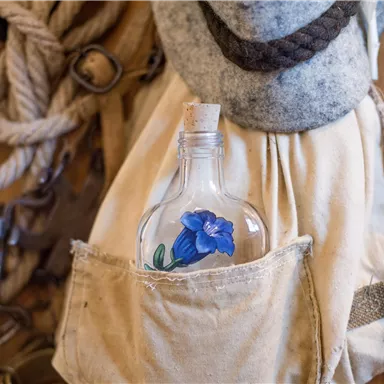 Eine Flasche mit einer blauen Blume steht in einer Tasche. Im Hintergrund sind Seile und weitere maritime Gegenstände zu sehen.
