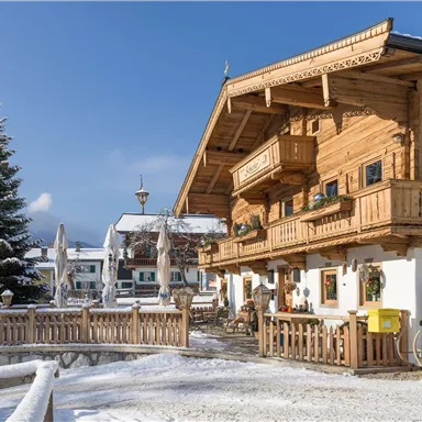 Ein gemütliches Holzhaus im Schnee mit Balkonen und einer schönen Terrasse. Im Hintergrund sieht man weitere Gebäude und einen Tannenbaum.