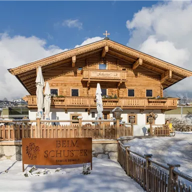 Ein traditionelles Holzhaus im Alpenstil, umgeben von Schnee. Im Vordergrund steht ein Schild mit der Aufschrift "Beim Schuster".