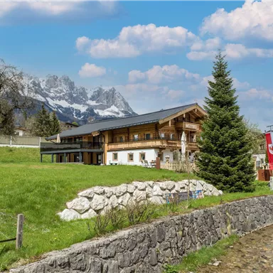 Eine gemütliche Berghütte umgeben von grünen Wiesen und Bäumen. Im Hintergrund sind beeindruckende Berge und ein klarer Himmel zu sehen.