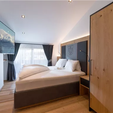 Ein modernes Schlafzimmer mit einem großen Bett und einem Fernseher an der Wand. Helle Farben und Holzdetails schaffen eine gemütliche Atmosphäre.