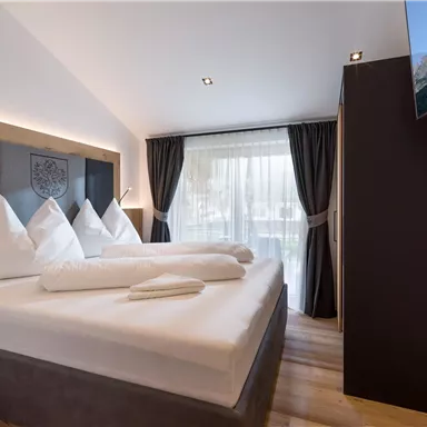 Ein modernes Hotelzimmer mit einem großen Doppelbett und eleganten Kissen. Große Fenster sorgen für viel Tageslicht und einen Ausblick nach außen.