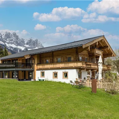 Ein schönes Holzhaus mit einem modernen Design, umgeben von grünen Wiesen. Im Hintergrund sind majestätische Berge und ein blauer Himmel zu sehen.