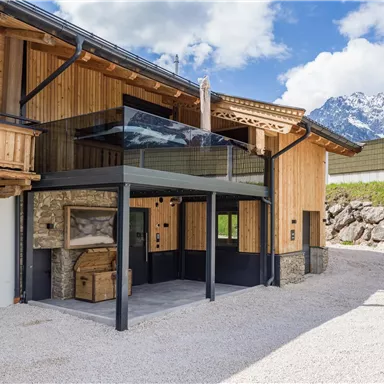 Ein modernes Chalet mit Holz- und Steinelementen in einer ländlichen Umgebung. Im Hintergrund sind Berge und ein blauer Himmel zu sehen.