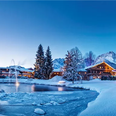 A picturesque winter landscape with snow-covered trees and a frozen pond. In the background, cozy cabins and mountains can be seen under a clear blue sky.