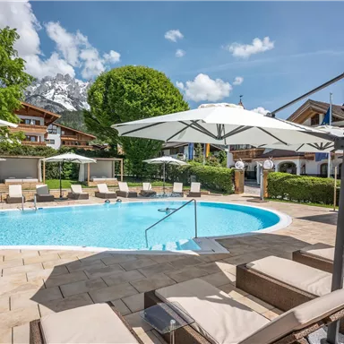 A beautiful pool area with loungers and sun umbrellas. In the background, there are green trees and charming architecture.