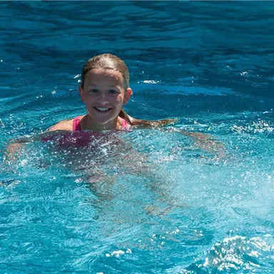 Ein Mädchen schwimmt fröhlich in klarem, blauem Wasser. Die Sonne scheint und zeichnet das Wasser mit Lichtpunkten.