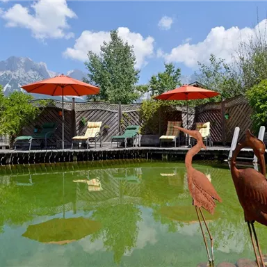 A quiet garden with a pond and two decorative flamingos. In the background, there are loungers and sun umbrellas under a blue sky.