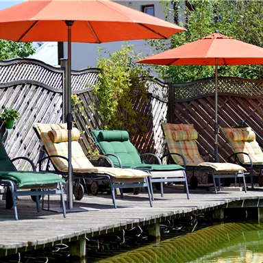 Eine gemütliche Terrasse mit Liegestühlen und Sonnenschirmen. Im Hintergrund sind Pflanzen und ein Zaun zu sehen.